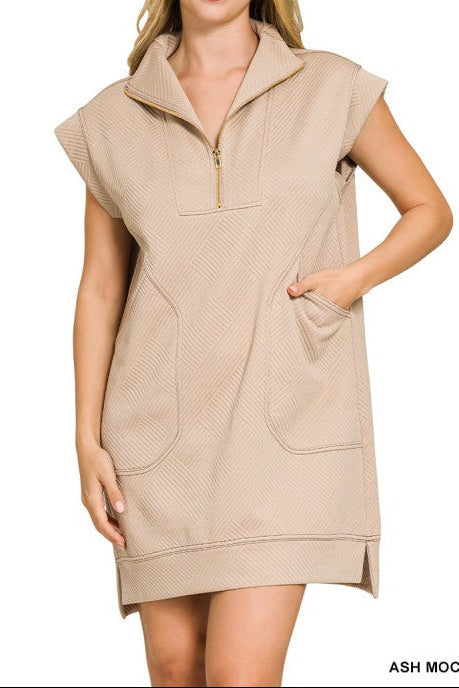 A woman modeling a sleeveless, half-zip mock-neck dress in a light beige fabric with a textured chevron pattern and large front patch pockets, set against a plain white background.
