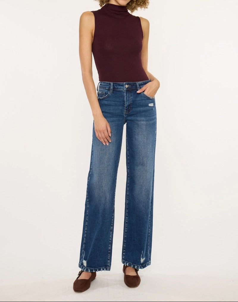A model with curly brown hair stands against a white studio background wearing a sleeveless dark burgundy mock-neck top and high-waisted, wide-leg blue jeans with brown flat shoes.