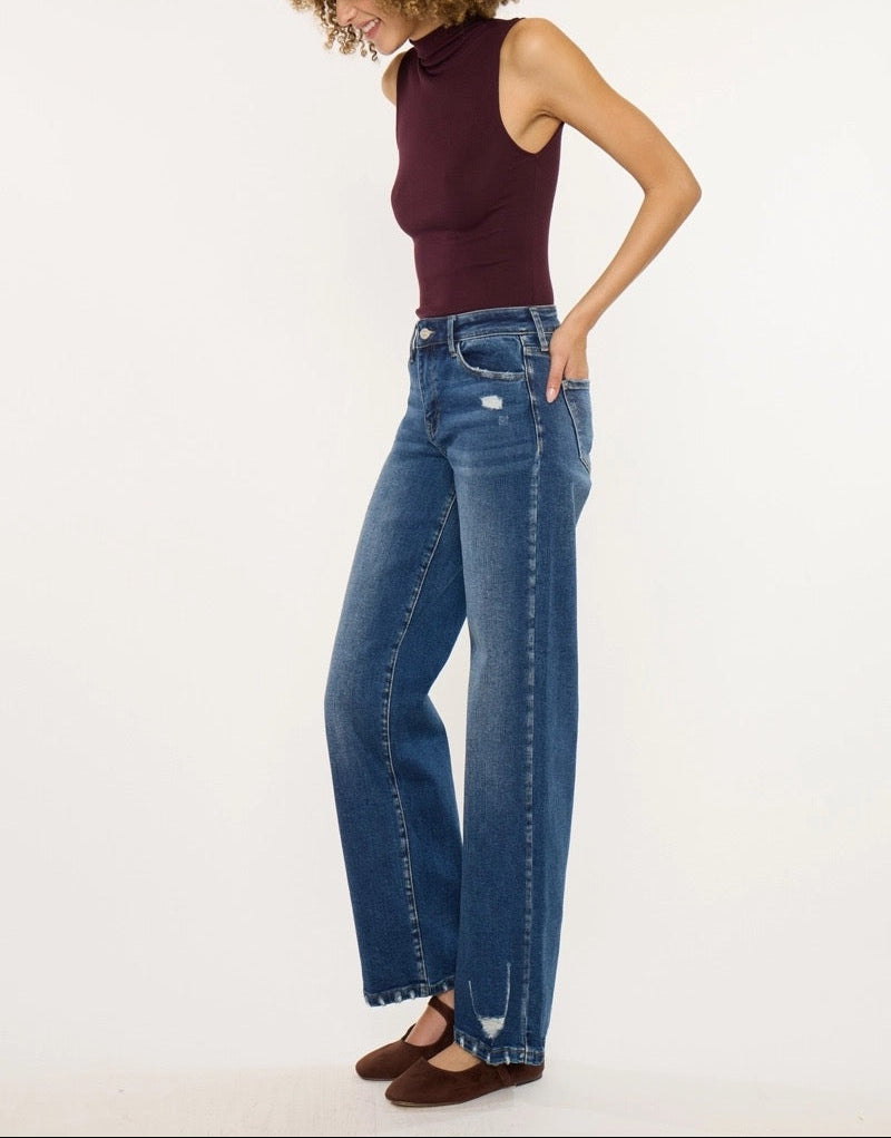 A woman modeling a pair of high-waisted, wide-leg blue jeans with a faded wash, paired with a sleeveless burgundy top and brown flats, set against a plain white studio background.