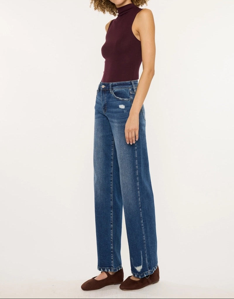 A woman modeling a pair of wide-leg blue jeans with a small distressed detail near the hem, paired with a sleeveless burgundy top and brown flats, against a plain white studio background.