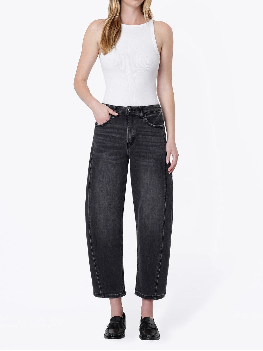 A woman modeling a pair of wide-leg cropped black jeans with a white sleeveless top and black loafers, standing against a plain white studio background.