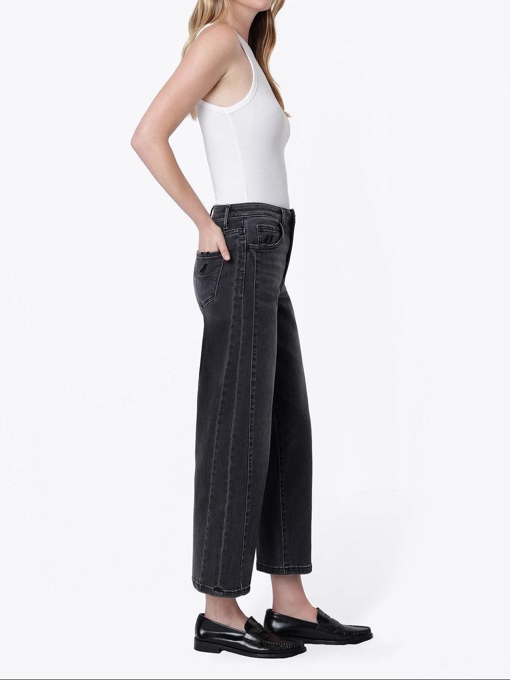 A woman stands in profile wearing a white sleeveless top, dark charcoal wide-leg cropped jeans, and black loafers against a plain white studio background.