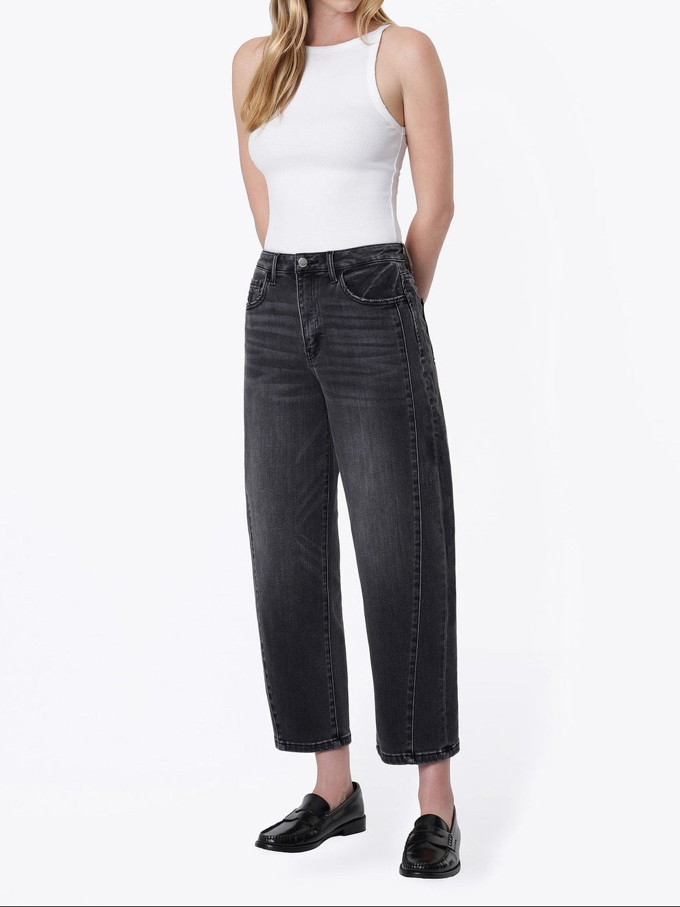 A woman modeling a pair of wide-leg cropped black jeans with a faded wash, paired with a white ribbed tank top and black loafers, against a plain white studio background.