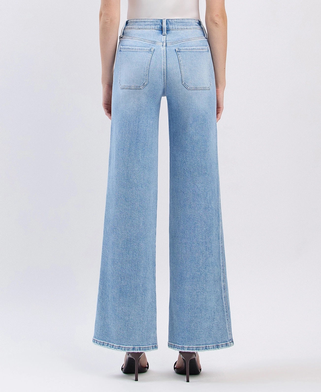 A person shown from the back wearing light-wash wide-leg denim jeans with a white top and black pointed heels, photographed against a clean light gray studio background.