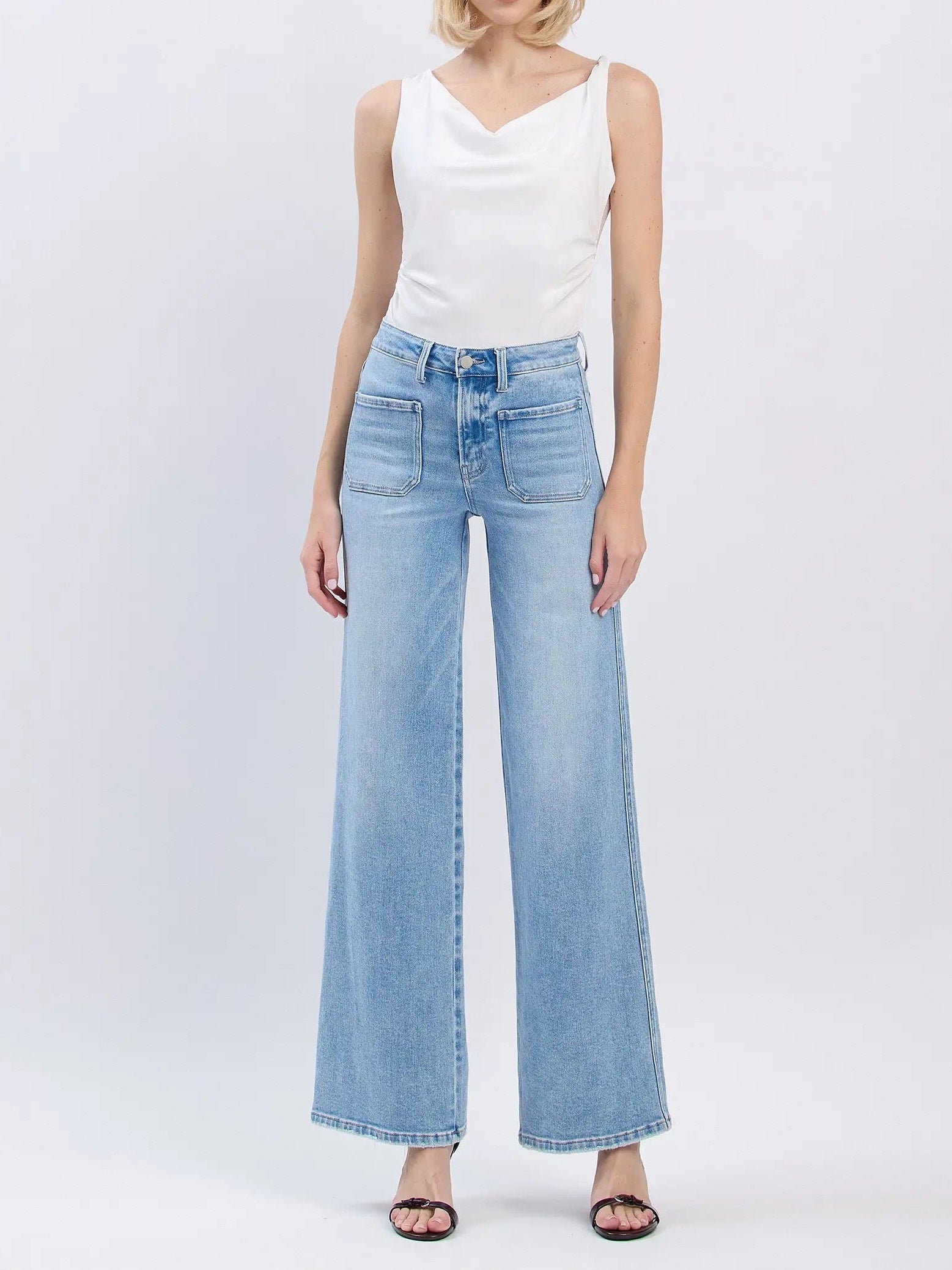 A woman modeling light-wash wide-leg denim jeans with a white sleeveless top, standing against a plain light gray studio background.