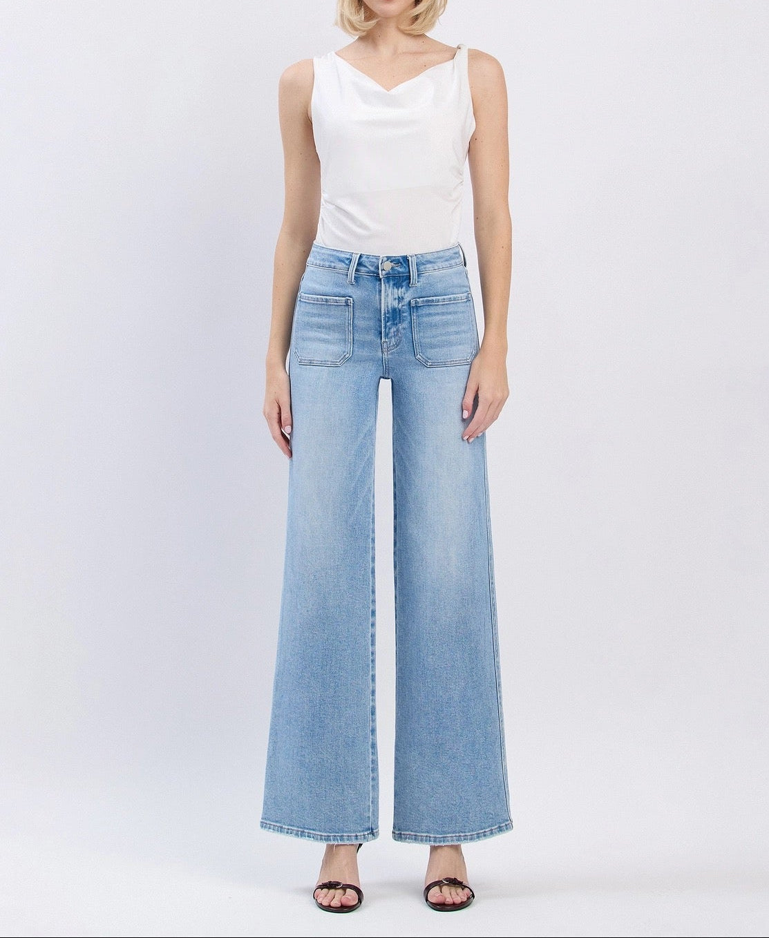 A woman stands against a light gray studio background wearing a white sleeveless top and light-wash wide-leg denim jeans with dark flat sandals.
