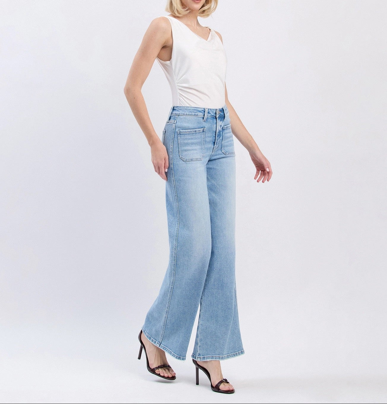 A woman modeling light-wash blue wide-leg jeans with a white sleeveless top and black heeled sandals, standing against a plain light gray studio background.