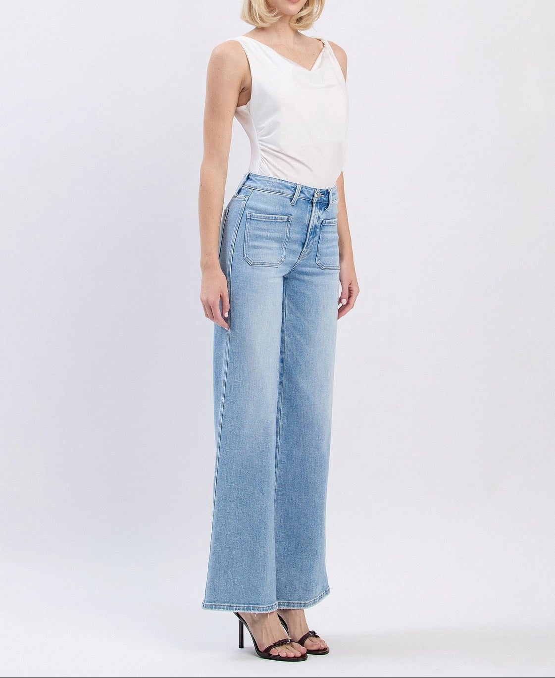 A woman modeling a pair of light-wash wide-leg denim jeans with a white sleeveless top and black heeled sandals, standing against a plain light gray studio background.