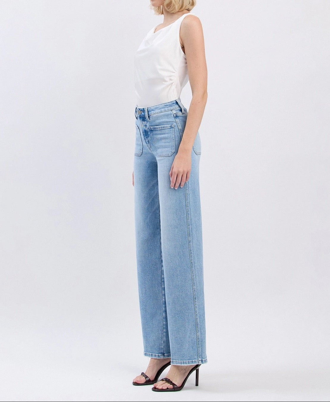 A woman stands in profile wearing light-wash wide-leg jeans, a white sleeveless top, and black heeled sandals against a plain light gray studio background.