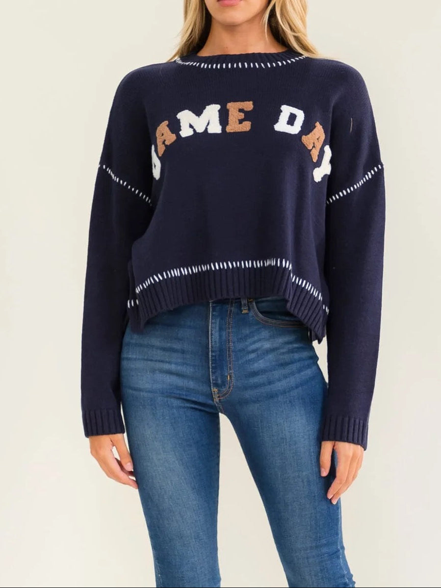 A woman modeling a navy blue knit sweater with 'GAME DAY' lettering across the chest in white, gold, and navy, paired with blue jeans against a plain light beige background.