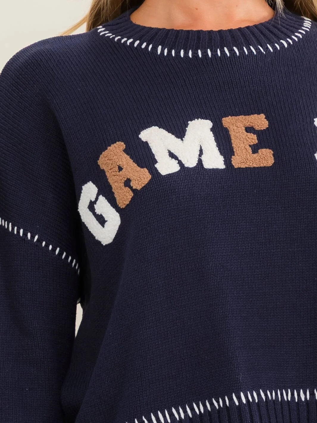 Close-up of a navy blue knit sweater with 'GAME' lettering across the chest in white and tan, plus white dashed stitch accents around the neckline and sleeves.