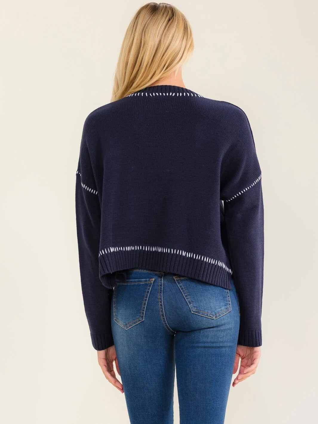 A woman viewed from the back wearing a navy blue knit sweater with white dotted trim details and blue jeans, set against a plain light beige background.