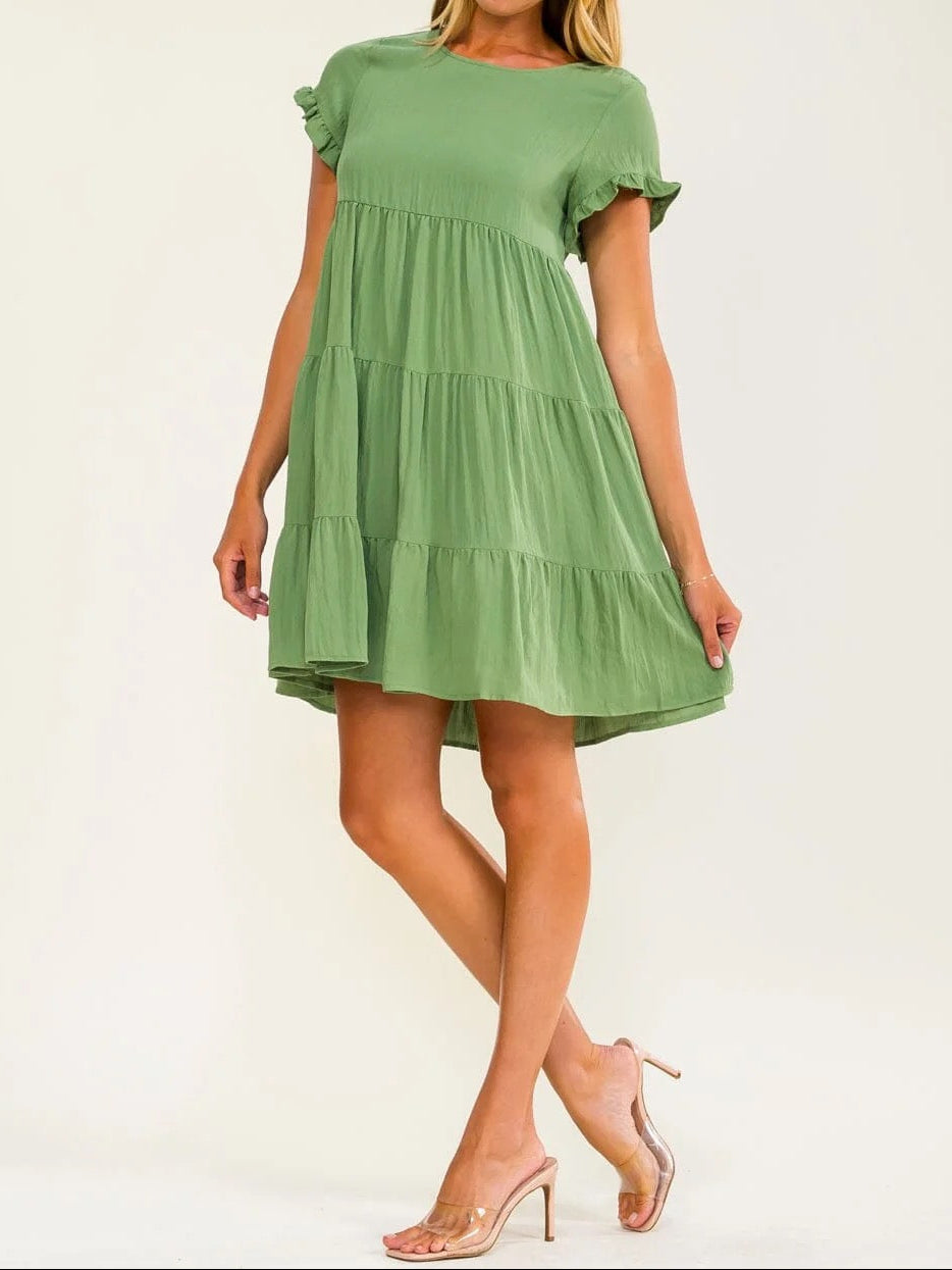 A woman modeling a sage green tiered mini dress with short flutter sleeves and ruffled trim, paired with clear high-heeled sandals, against a plain off-white studio background.