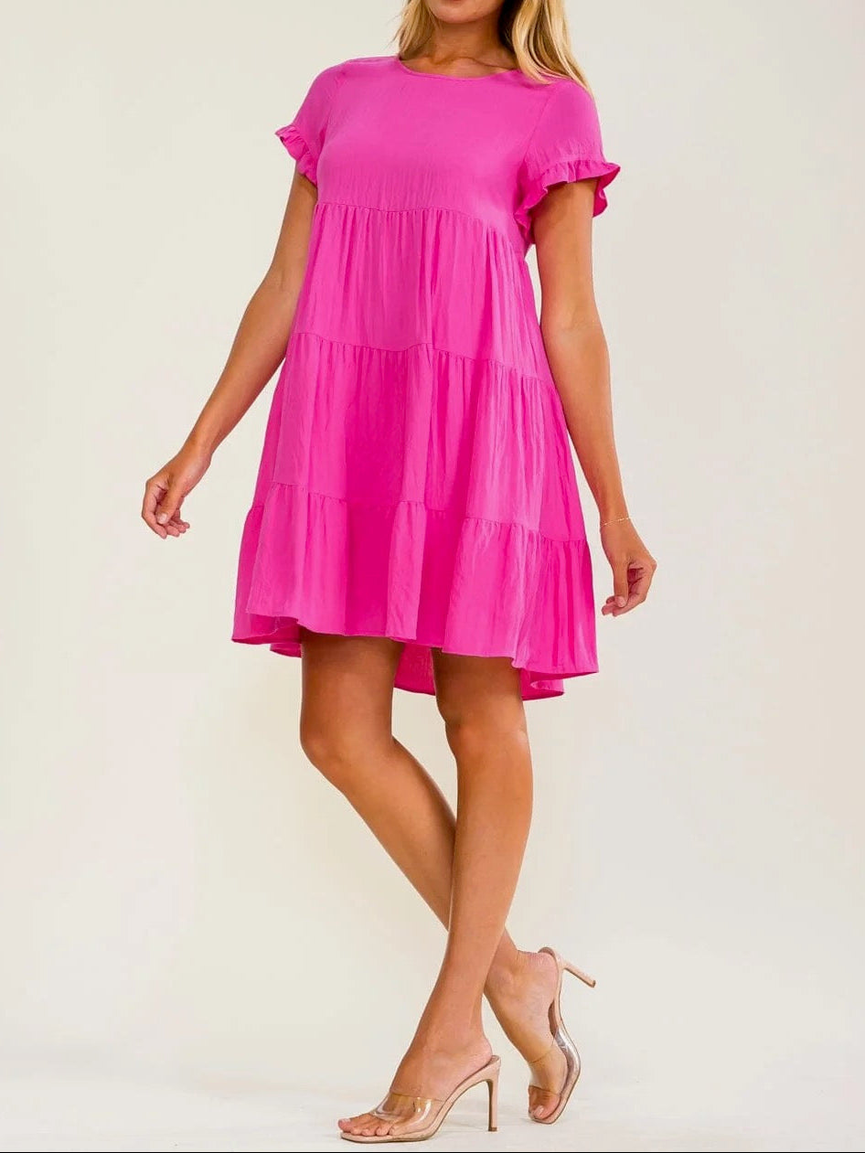 A woman modeling a bright pink tiered mini dress with short flutter sleeves and ruffled trim, paired with nude high-heeled sandals, against a plain off-white studio background.