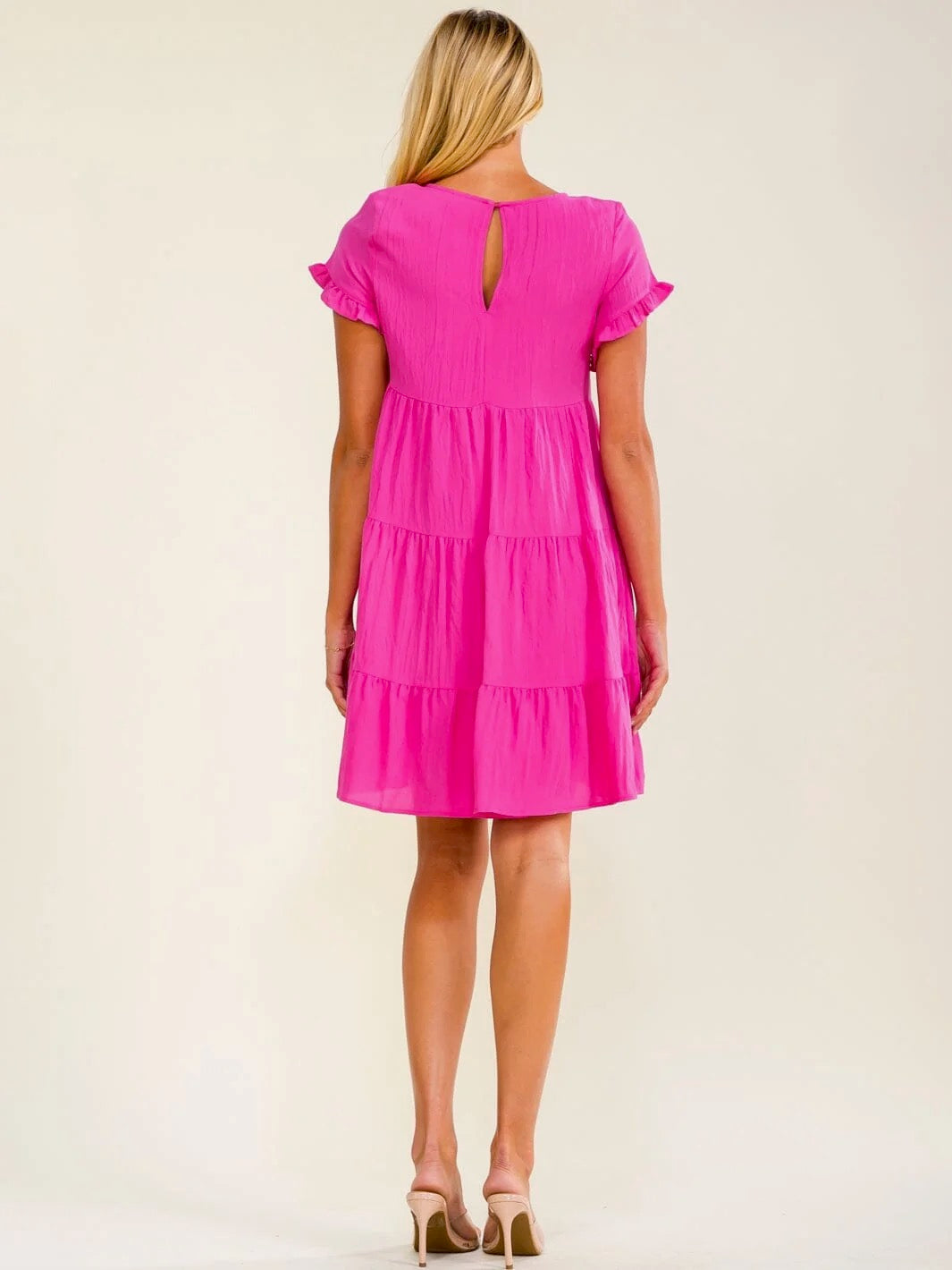 A woman viewed from the back wearing a bright pink tiered dress with short ruffled sleeves and a small keyhole cutout at the back, paired with beige high heels against a plain light beige studio background.