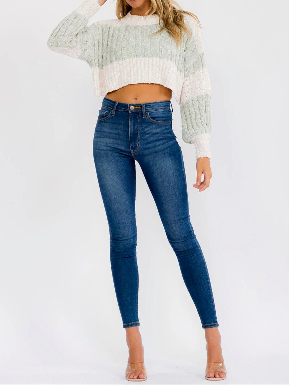 A woman modeling a light sage green and white striped cropped knit sweater with long balloon sleeves, paired with dark blue skinny jeans and nude high-heeled sandals, against a plain white studio background.