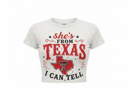 A white cropped short-sleeve t-shirt featuring a red and black graphic print that reads 'she's FROM TEXAS I CAN TELL' with a Texas-shaped outline and 'T' logo in the center, displayed on a clean white background.